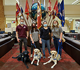 One male and three female college students stand in front of flags and a globe with one black and two yellow Labradors in yellow vests.