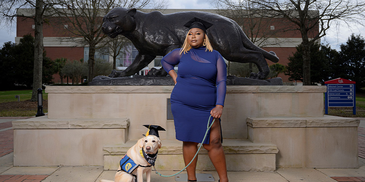 A woman with dark hair and a red shirt standing and holding a leash attached to a white Labrador service dog sitting next to her