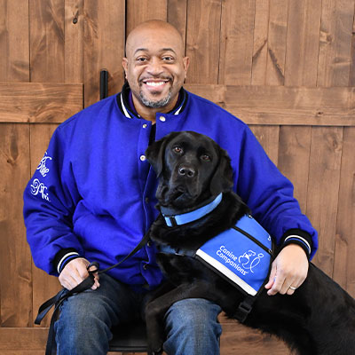 A young puppy in a service dog cape looks up to an adult labrador in a service dog vest