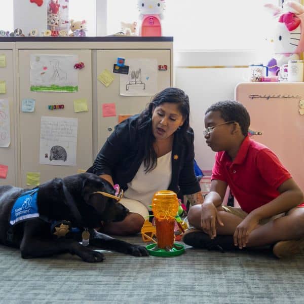 a woman sitting down on the floor with a child and a Labrador; dog is wearing sunglasses as he lays down on the carpet; dog is wearing a blue vest