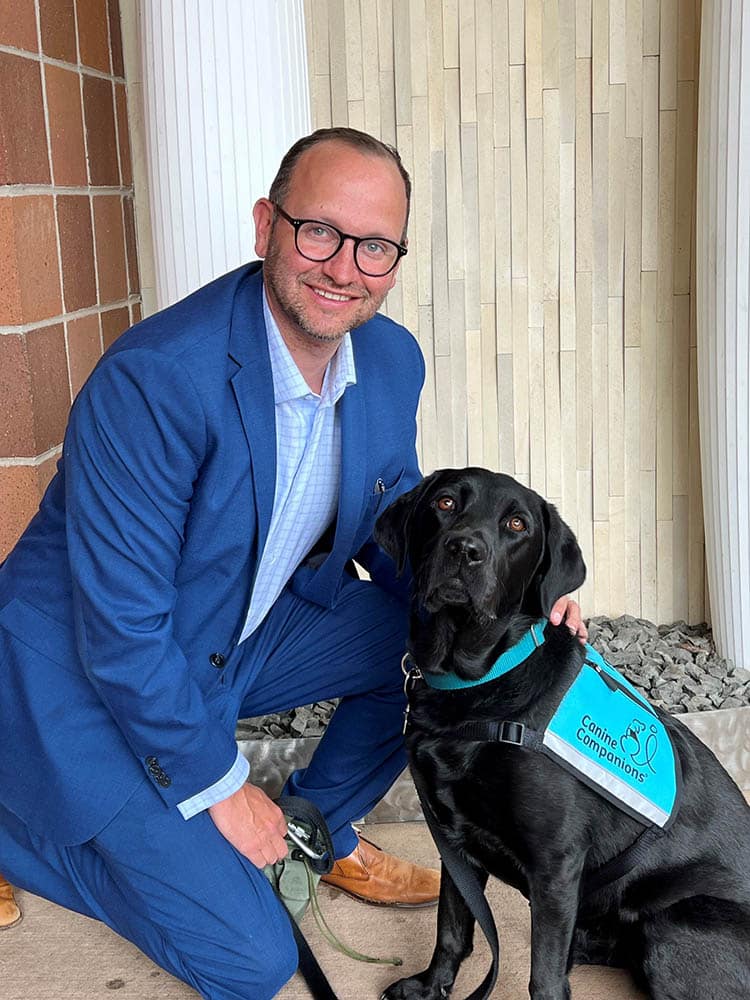 a smiling man a in a suit with his arm around a black lab in a teal therapy dog vest