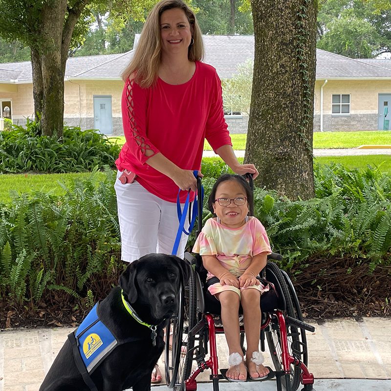 A young Asian girl in a wheelchair smile with her parent and a black Labrador service dog