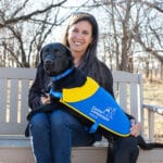 A smiling woman sitting on a bench outdoors with a black lab in a canine companions matriculation cape laying across her lap