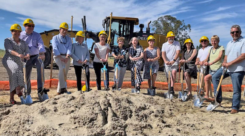 a row of people in yellow hardhats stand with shovels in front of a backhoe