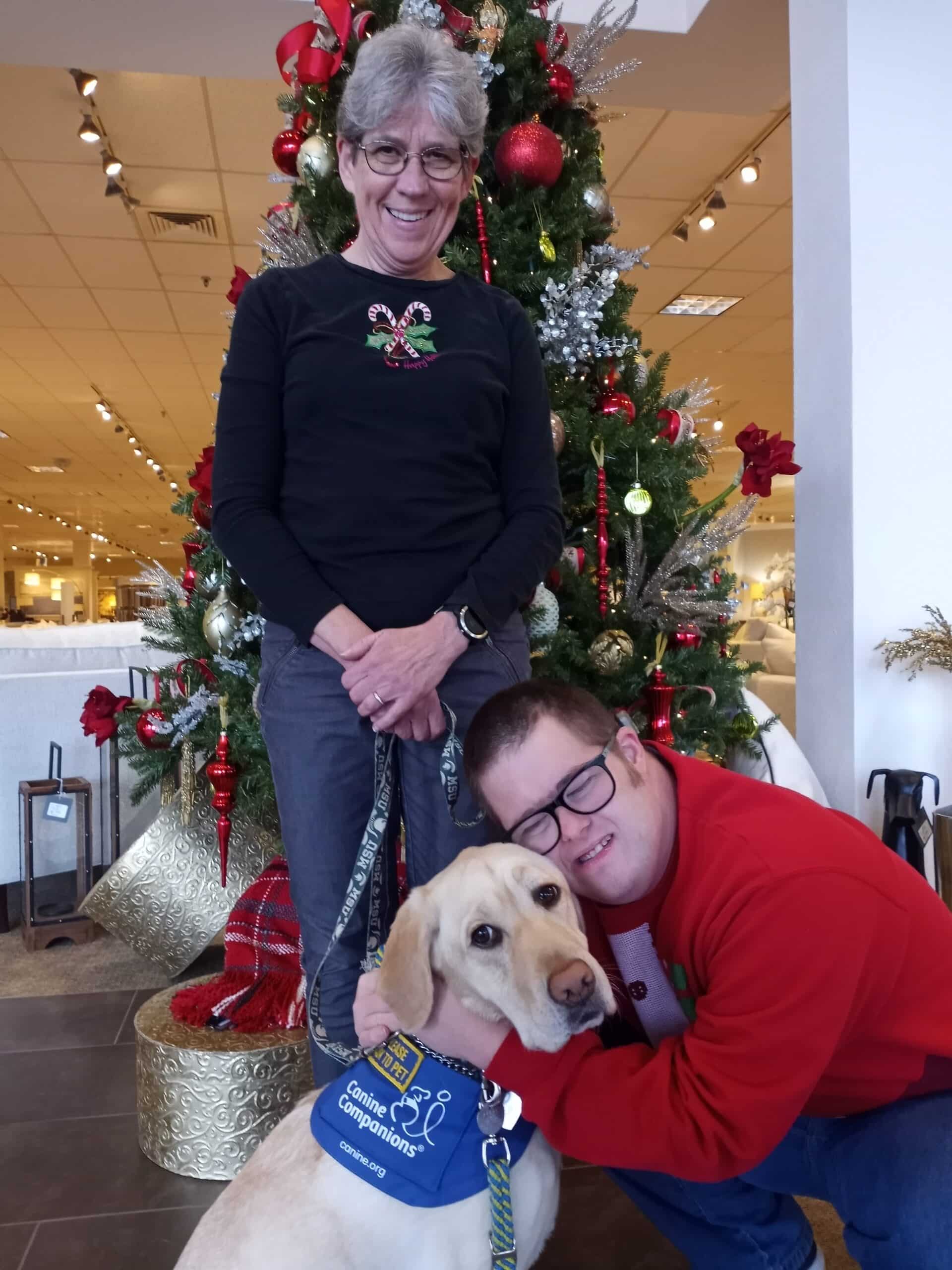 A  young man hugs his service dog with his mother standing behind