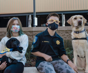 13 year old Miller and his yellow service dog, Bahama, sit with his younger sister Jac holding black Canine Companions puppy O'Sully