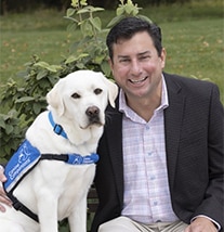 A smiling man sitting next to a yellow lab wearing a blue canine companions vest