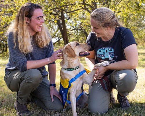 two young volunteers next to a yellow lab puppy in a yellow puppy vest