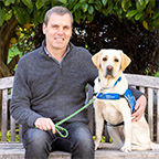 Image of Keith Edwards sitting on a bench with his arm around a yellow lab in a blue service vest sitting beside him