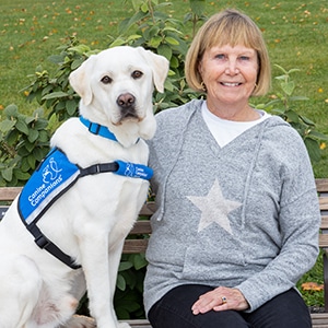 A smiling woman sitting next to a yellow lab service dog in a blue canine companions vest