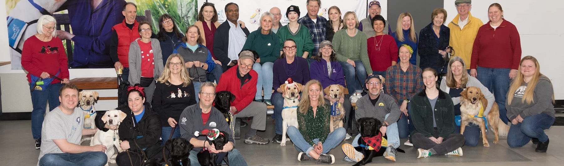 A large group of smiling volunteers with canine companions puppies in yellow puppy vests