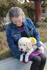 Rachel Meyer sits looking at a yellow Canine Companions puppy on her lap