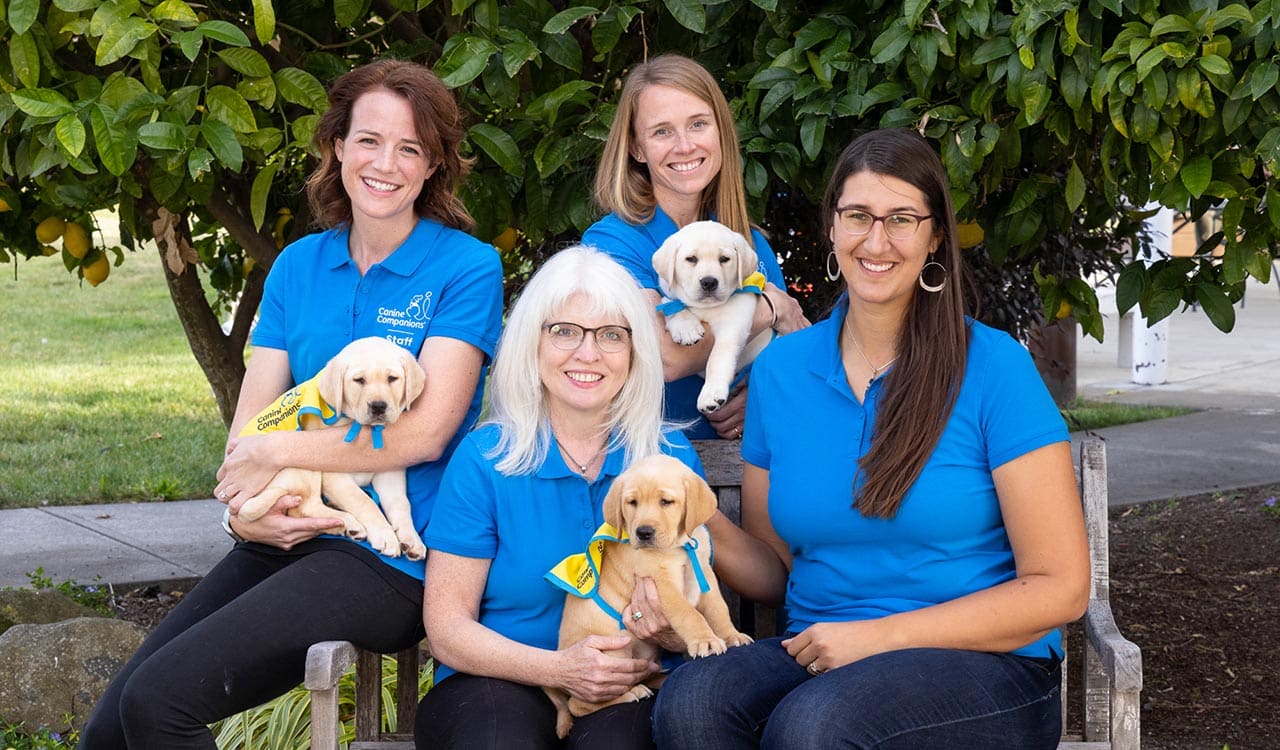 Four women from the Canine Companions science team sitting on a bench holding puppies