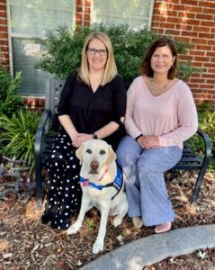 two smiling women sitting in front of a house with a yellow lab in a blue service vest