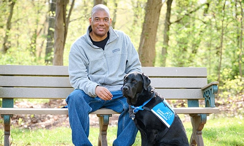 a man sitting on a bench with a black lab in a blue service vest sitting on the ground at his feet