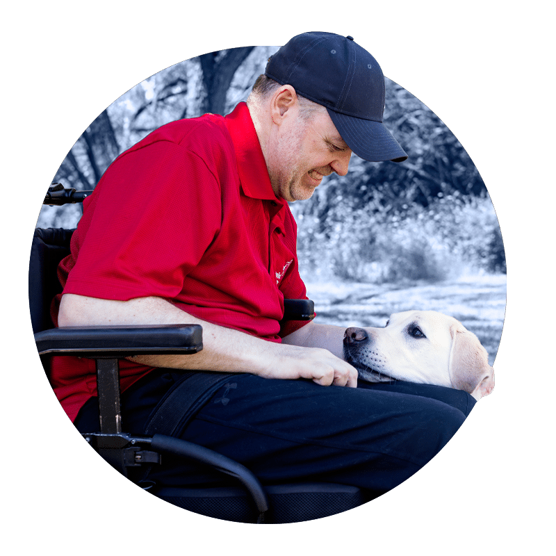 a smiling man in a wheelchair with a yellow lab in a blue service vest laying across his lap