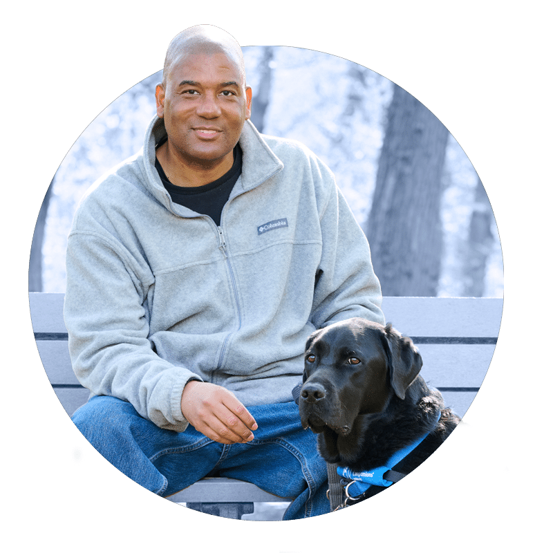 a man sitting on a bench with a black lab in a blue service vest sitting on the ground at his feet